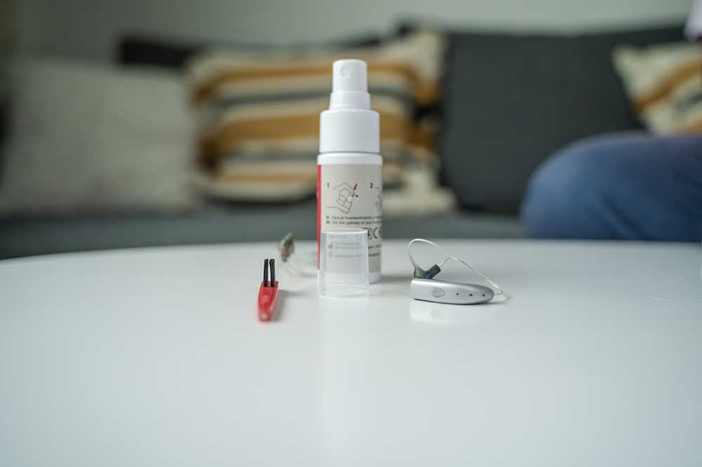 Cleaning spray, a red brush and a behind-the-ear hearing aid on a white table, hearing aid care tools from Port Elgin Audiology in Port Elgin, ON